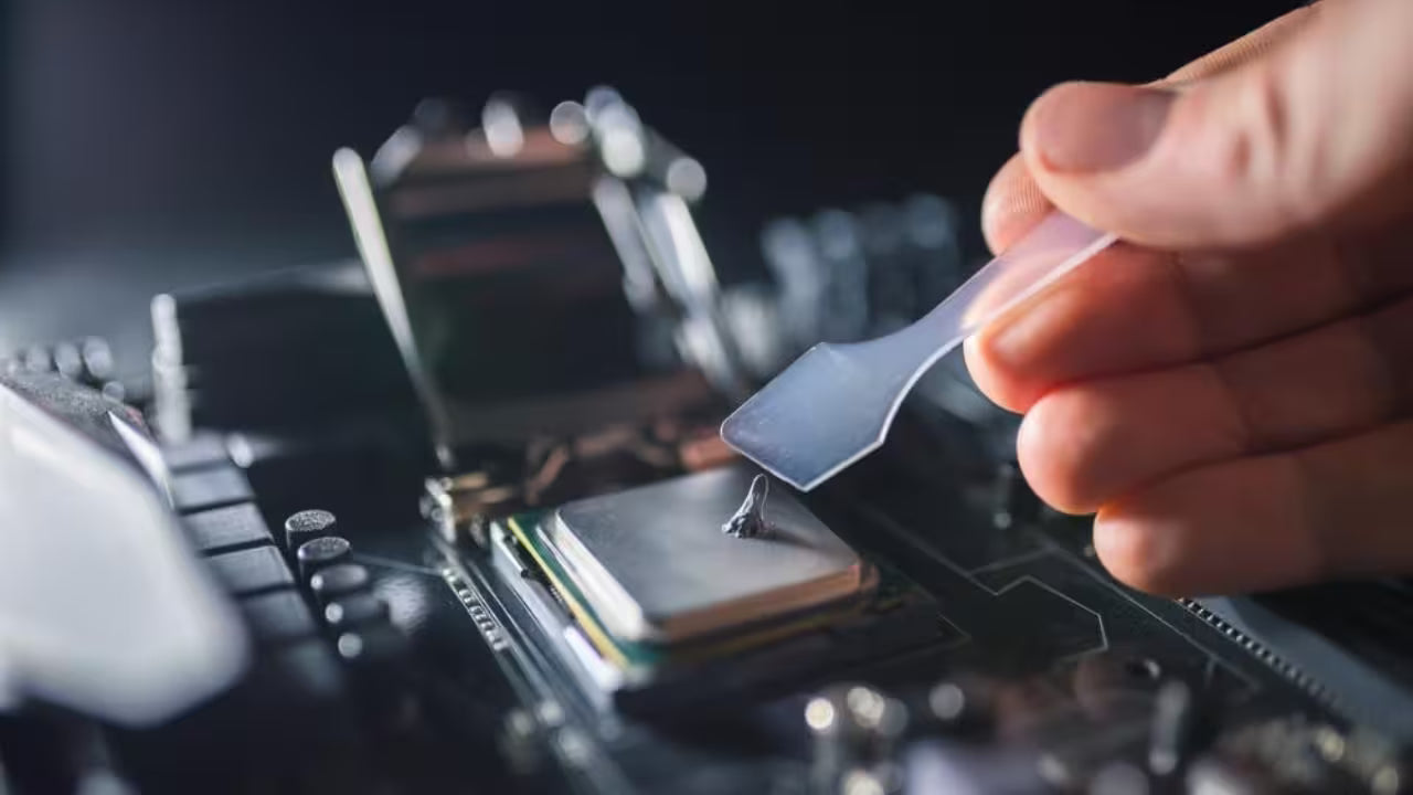 Close-up of a person using a small plastic spatula to spread thermal paste onto an Intel processor installed in a motherboard socket